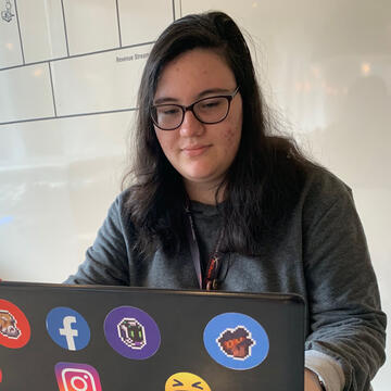 Foto de Carina. Garota branca de cabelos longos e pretos, sentada enquanto usa um notebook com adesivos.